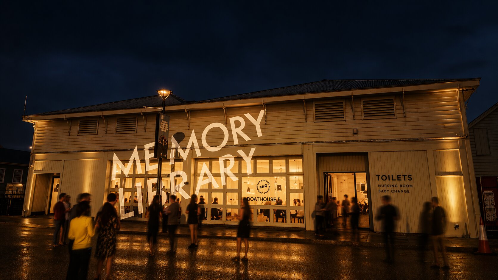Boathouse 5 at night, with the Memory Library wordmark projected across the front of the building and visitors gathered outside.