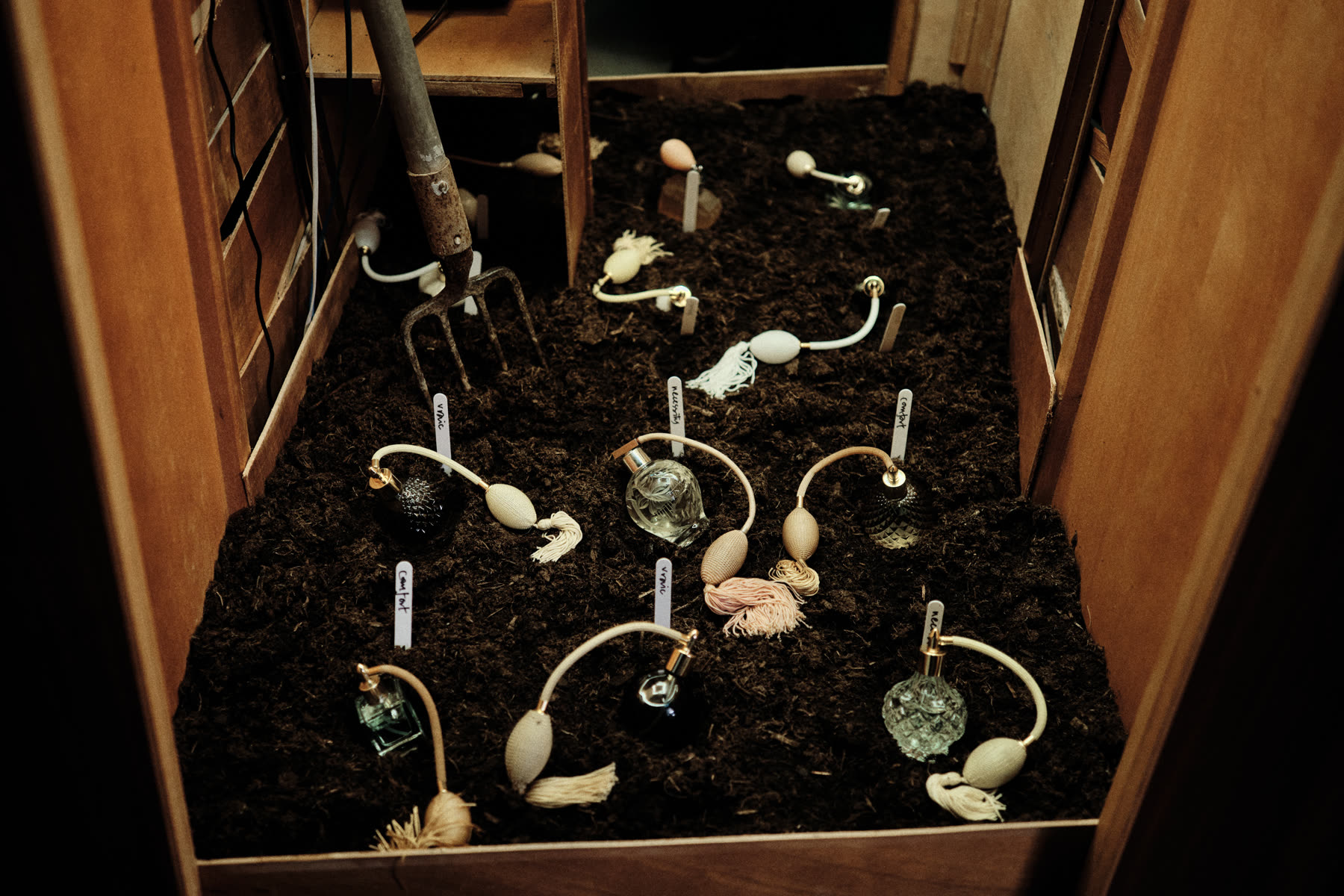Vintage perfume atomisers planted in soil, each labelled by hand with a remembered scent.