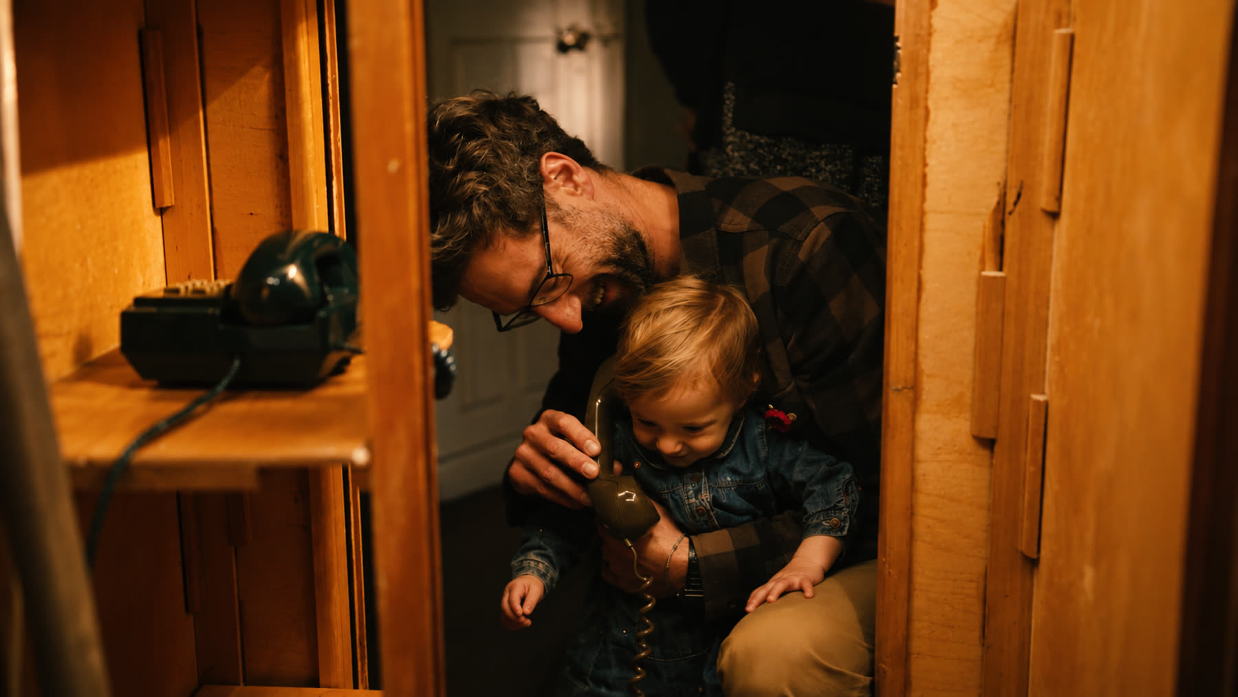 A man helps a young child hold an old rotary telephone receiver inside a wooden booth-like sculpture.